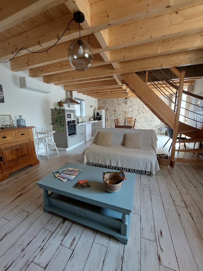 A bright living area features wooden beams and a light wood floor. A spacious sofa with two cushions faces a low table, adorned with magazines and a decorative basket. The kitchen is visible in the background, with modern appliances and rustic elements.