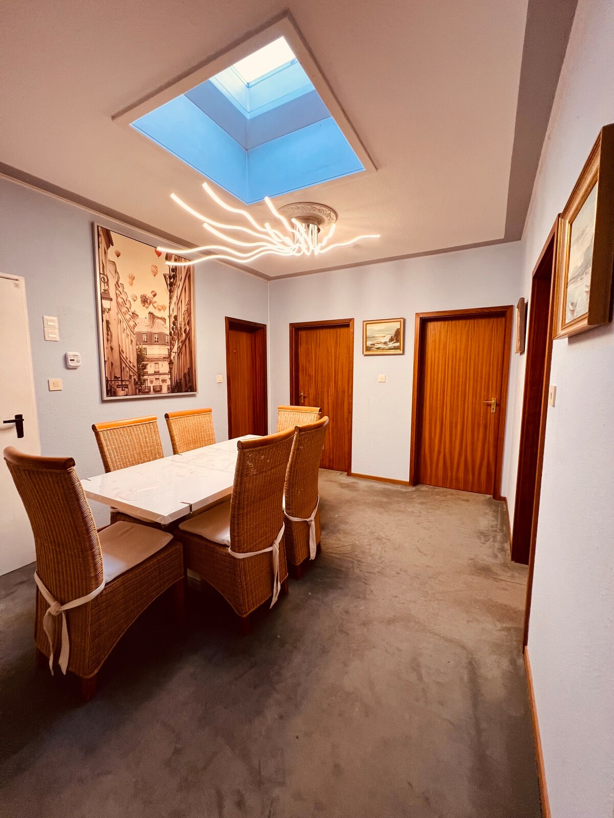 A dining area features a large table surrounded by six rattan chairs. Soft gray carpeting extends across the floor. A statement light fixture hangs above the table, and natural light enters through a skylight. Four wooden doors line the walls, leading to additional rooms.