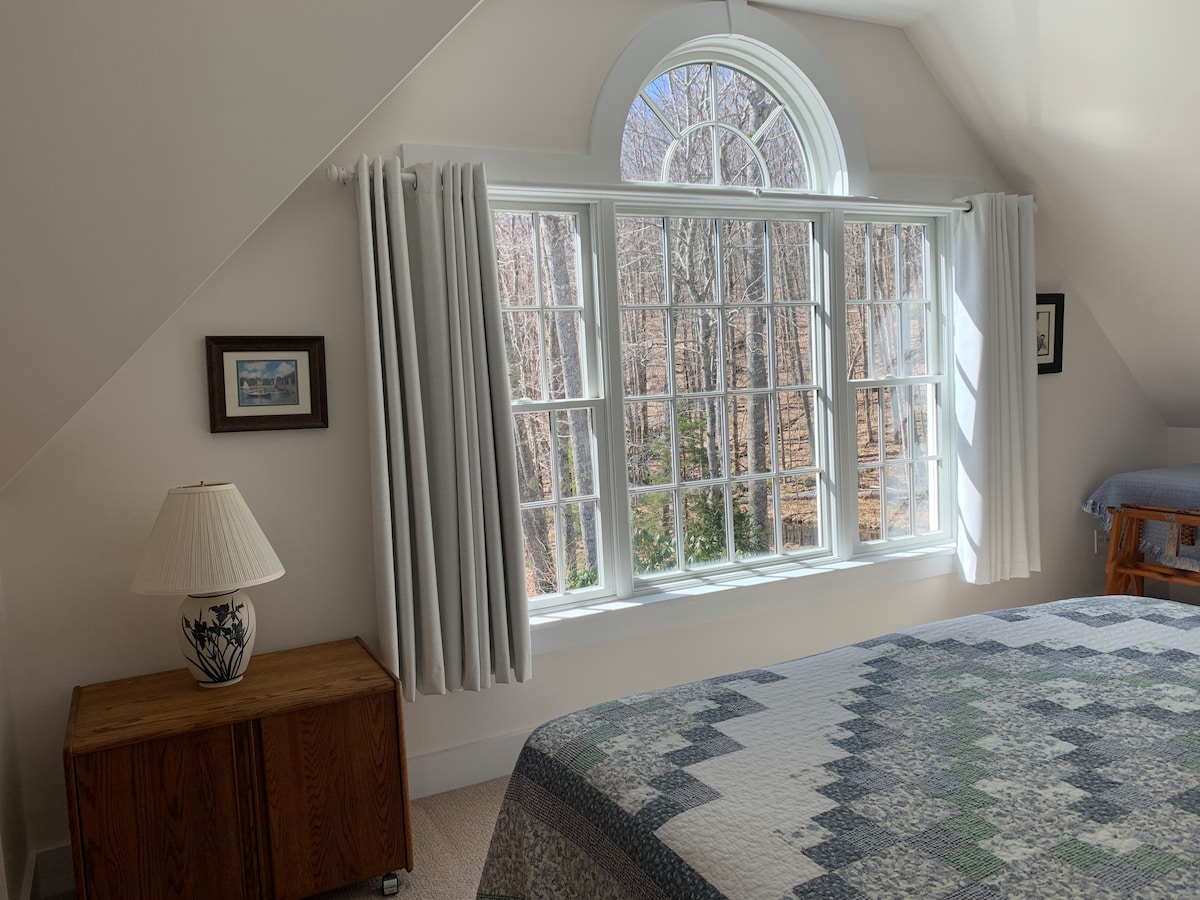 A bright bedroom is presented with a spacious bed covered in a quilt, accompanied by a wooden cabinet. Natural light enters through a large arching window adorned with white curtains, revealing views of the surrounding woods, while framed artwork adds a touch of charm.