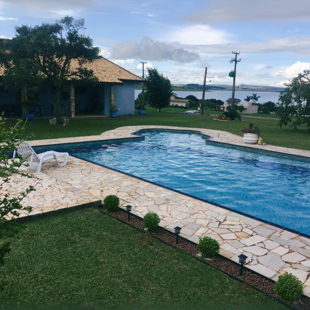 A bright pool features crystal-clear water surrounded by a stone patio. Lush green grass borders the area, and a few small hedges line the path. In the background, a house with a tiled roof can be seen, along with a scenic view of the water.