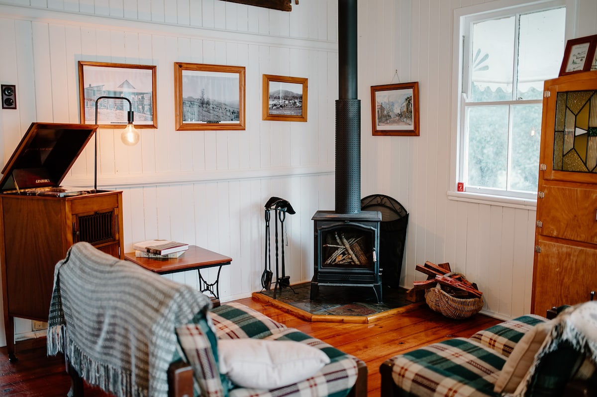 A cozy living area features plaid sofas and a wooden table beside a wood-burning stove. Black-and-white photographs hang on the walls, and a vintage record player stands next to the window, allowing for natural light to brighten the space.
