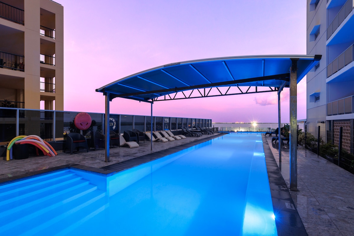 An infinity pool is featured, with a spacious deck area equipped with lounge chairs under a shaded canopy. The calm water reflects the pastel colors of the twilight sky, creating a serene ambiance suitable for relaxation.