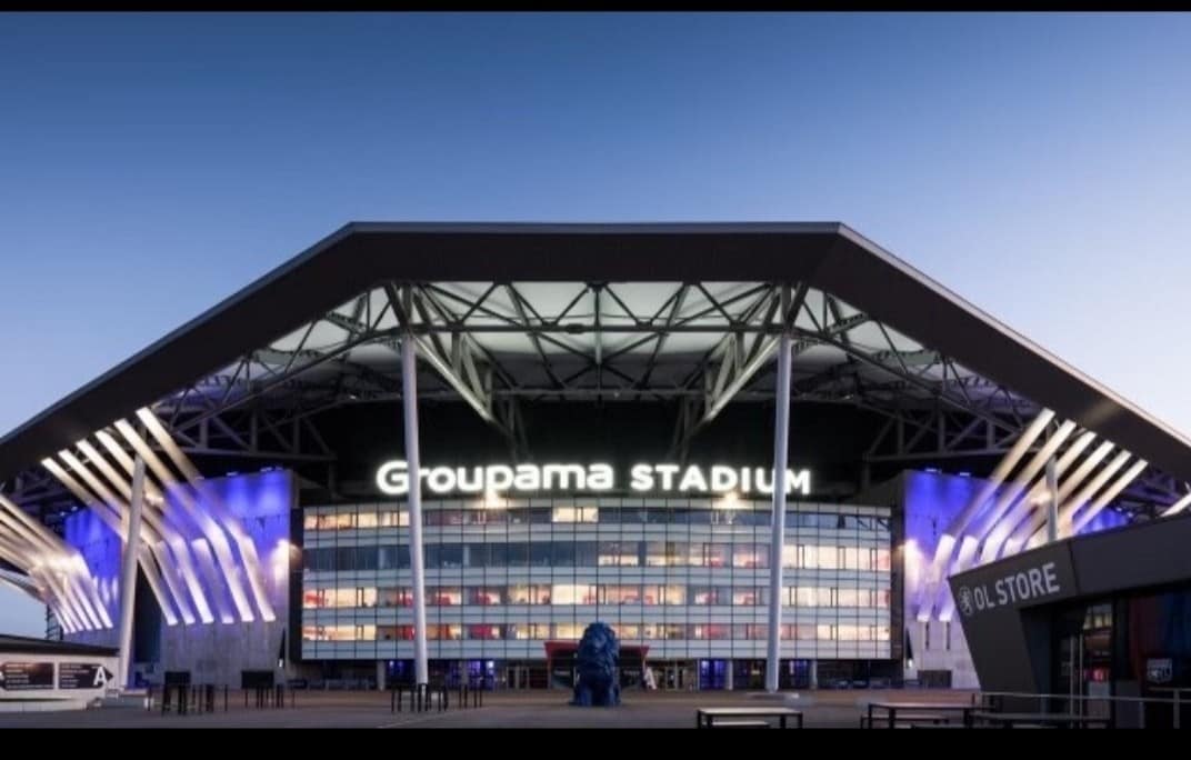 The facade of Groupama Stadium is highlighted by modern architecture and a distinctive roof structure. The stadium's name is prominently displayed in illuminated letters, set against a backdrop of evening sky. Nearby, a retail store entrance is visible, adding to the vibrant atmosphere.