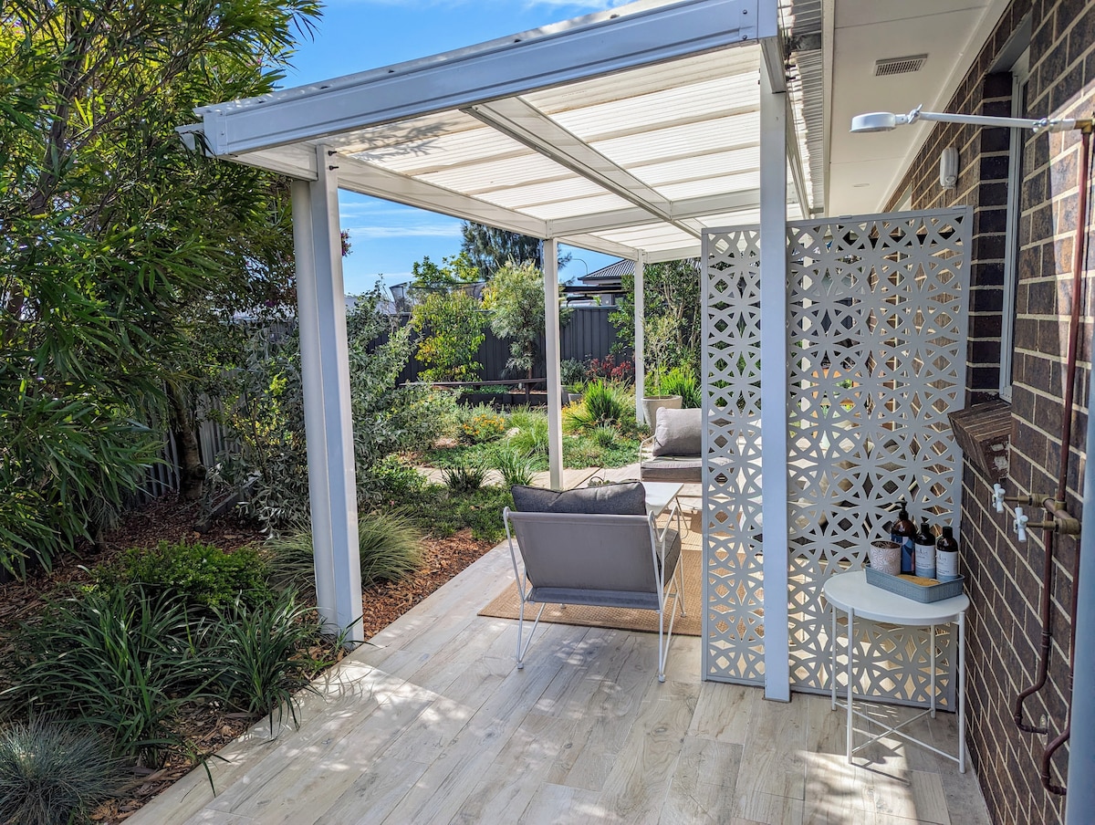 An outdoor entertaining area is framed by a covered verandah, featuring comfortable seating and a small side table. Lush greenery surrounds the space, while a decorative privacy screen adds texture and visual interest. Natural light enhances the inviting atmosphere.