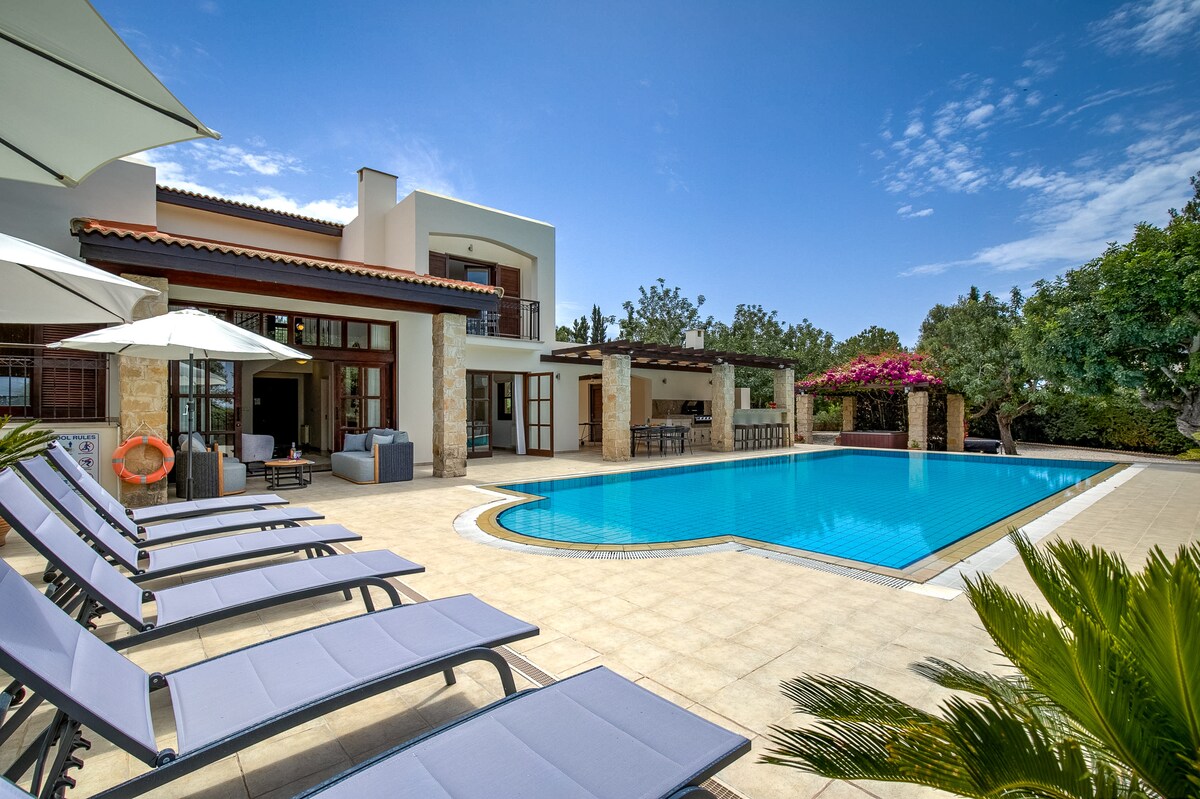 A spacious outdoor area showcases a private pool bordered by sunbeds. The pool is framed by a large patio, featuring a covered terrace with seating and an outdoor dining area. Lush greenery and flowering plants add touches of color around the garden.