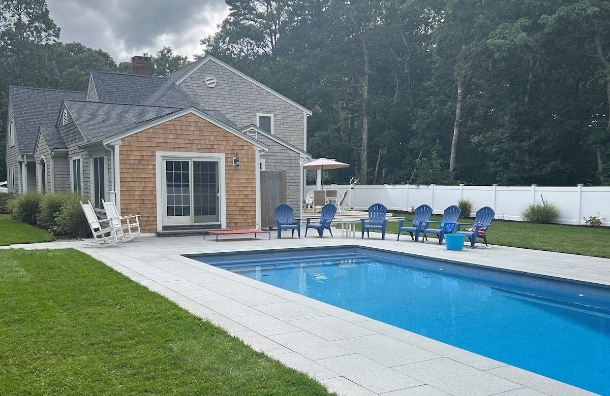 A spacious outdoor area features a refreshing pool bordered by a smooth stone patio. Blue lounge chairs are arranged around the pool, and the house is seen in the background, showcasing a blend of shingle and clapboard siding. Lush greenery adds a natural touch.