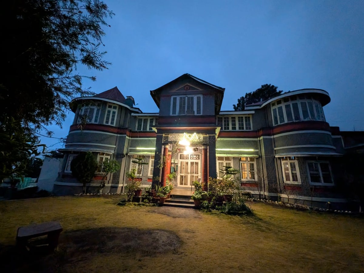 The grand heritage villa is illuminated softly at dusk, showcasing its colonial architecture with large windows and distinct curved balconies. Lush greenery surrounds the entrance, enhancing the inviting ambiance of the property, while a warm light highlights the detailed facade.