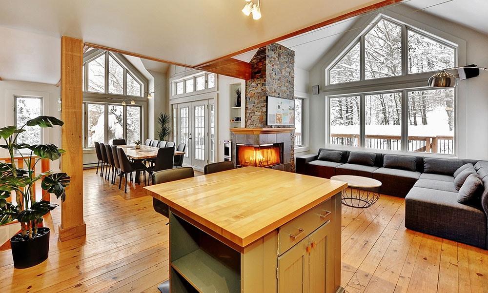 An open living space features large windows revealing snowy outdoor views. A dining area with a wooden table accommodates multiple guests, while a spacious sectional sofa is situated near a stone fireplace. Light wood flooring complements the welcoming atmosphere of the chalet.