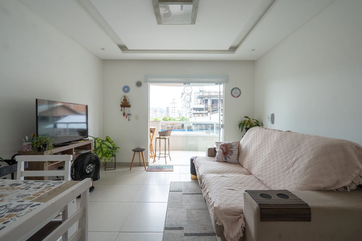 A bright living area features a cozy sofa covered with a light blanket, positioned near a window that allows natural light to fill the space. A television is present, and a small dining table with two chairs is visible, creating an inviting space for relaxation.