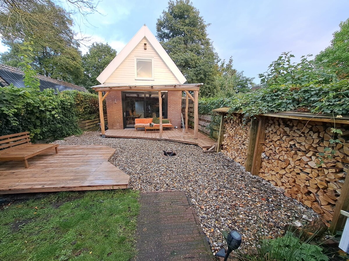 The exterior of the garden retreat features a wooden deck with seating and a natural stone pathway. Lush greenery surrounds the property, providing privacy. A stack of firewood is neatly arranged beside the structure, enhancing the rustic atmosphere.