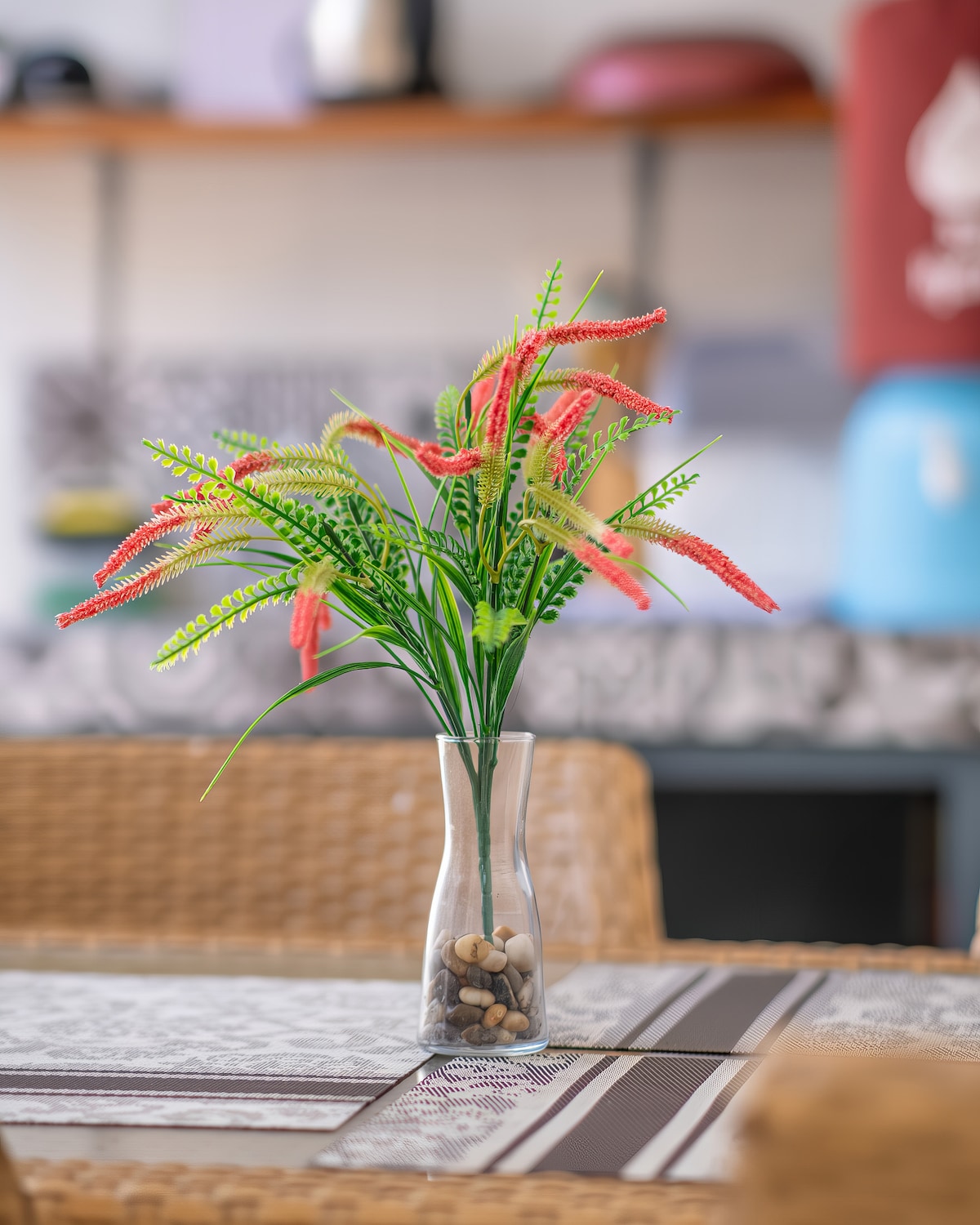 A decorative arrangement of artificial flowers is displayed in a clear glass vase filled with small stones. The vase sits on a dining table, enhancing the subtle decor in the background, which features a kitchen area with wooden accents.