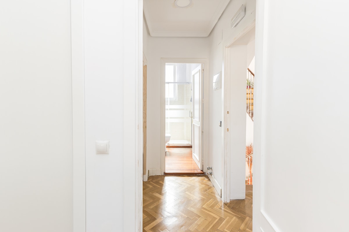 A hallway with light-colored walls and polished wooden flooring extends towards an open space at the end. Natural light is diffused through a window, illuminating the area. A doorway on the left leads to a separate room, contributing to the airy feel.