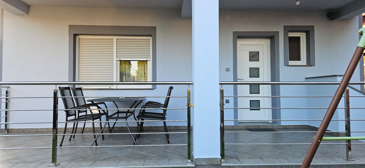 A private terrace features a metal dining table with four chairs, providing a space for outdoor relaxation. The entrance to the studio apartment is visible, along with a large window, allowing natural light to brighten the area.