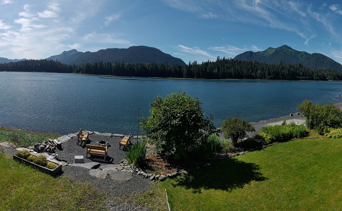 A panoramic view of the waterfront is displayed, featuring a serene waterway bordered by lush greenery and distant mountains. A seating area with benches is positioned on a stone patio, offering a peaceful spot to enjoy the natural surroundings.