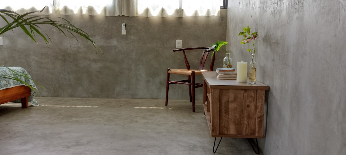 A minimalist interior features concrete walls, enhancing the spaciousness of the room. A wooden bench with decorative items sits beside a chair, while soft natural light filters through the sheer window coverings, creating a calm and serene environment.