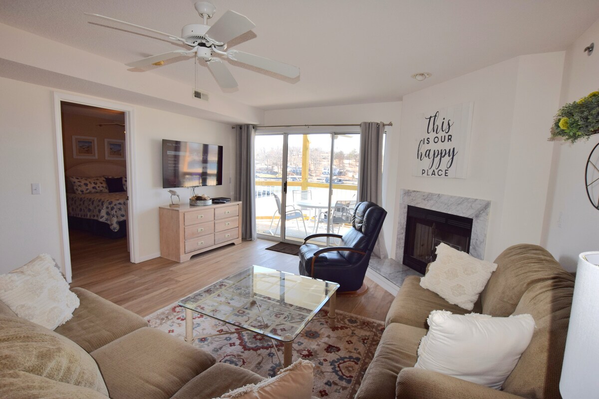 A living area features a comfortable sofa and armchair, with a coffee table positioned at the center. A flat-screen TV is mounted on the wall, and a marble fireplace adds a cozy touch. Sliding glass doors lead to a balcony with views of the outdoors.