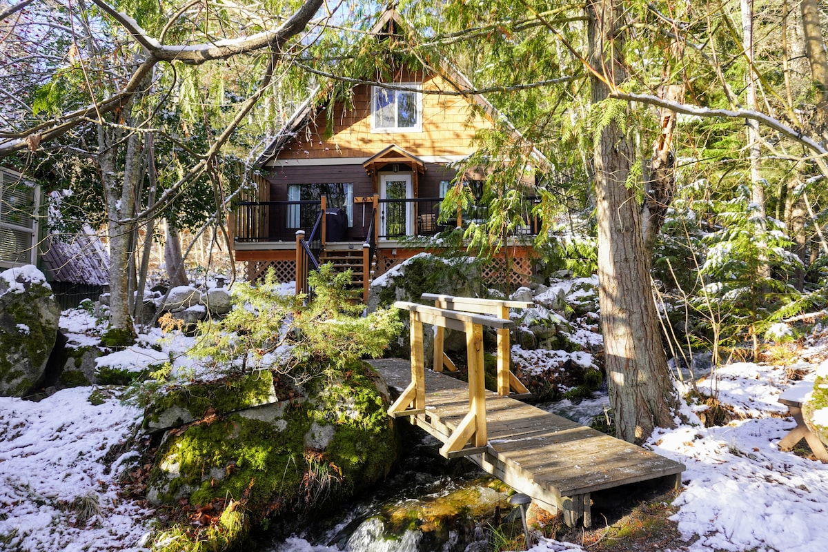 A wooden footbridge connects a lush, snow-dusted landscape to a charming log cabin. The cabin, set amid evergreen trees, features a welcoming porch with a set of chairs. Rocks and gentle stream flow are visible, enhancing the natural surroundings.