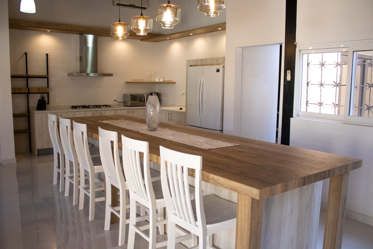A spacious kitchen is depicted, featuring a large wooden island with ample seating for eight. Modern pendant lights hang above, illuminating the area. A stainless steel refrigerator and a hooded stovetop are visible against the wall, complemented by sleek cabinetry and an inviting atmosphere.