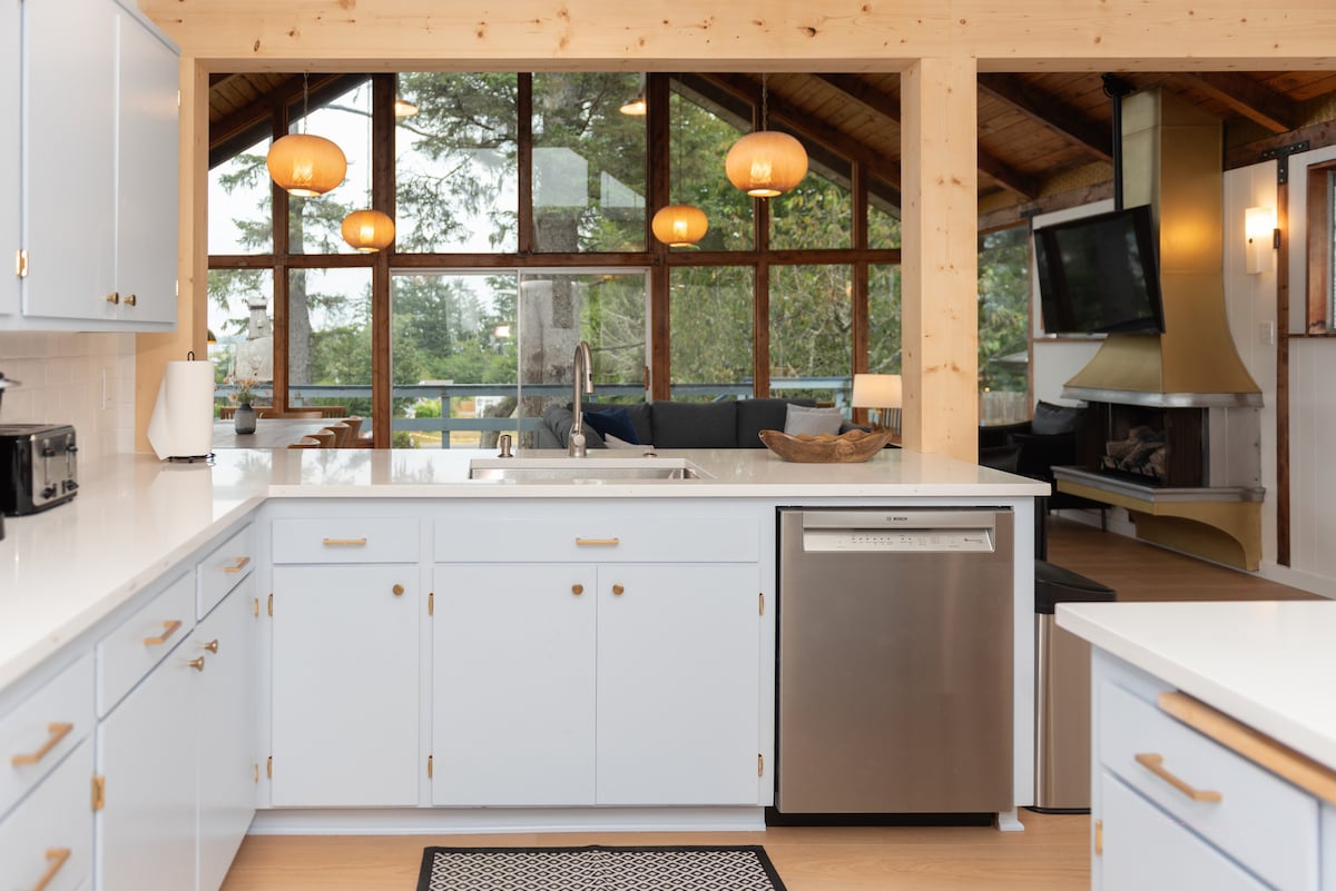 A bright kitchen is showcased with white cabinets and a large island. Above, pendant lights hang, illuminating the space. Large windows provide views of the surrounding greenery and natural light. A cozy living area, featuring a fireplace and seating, is visible in the background.