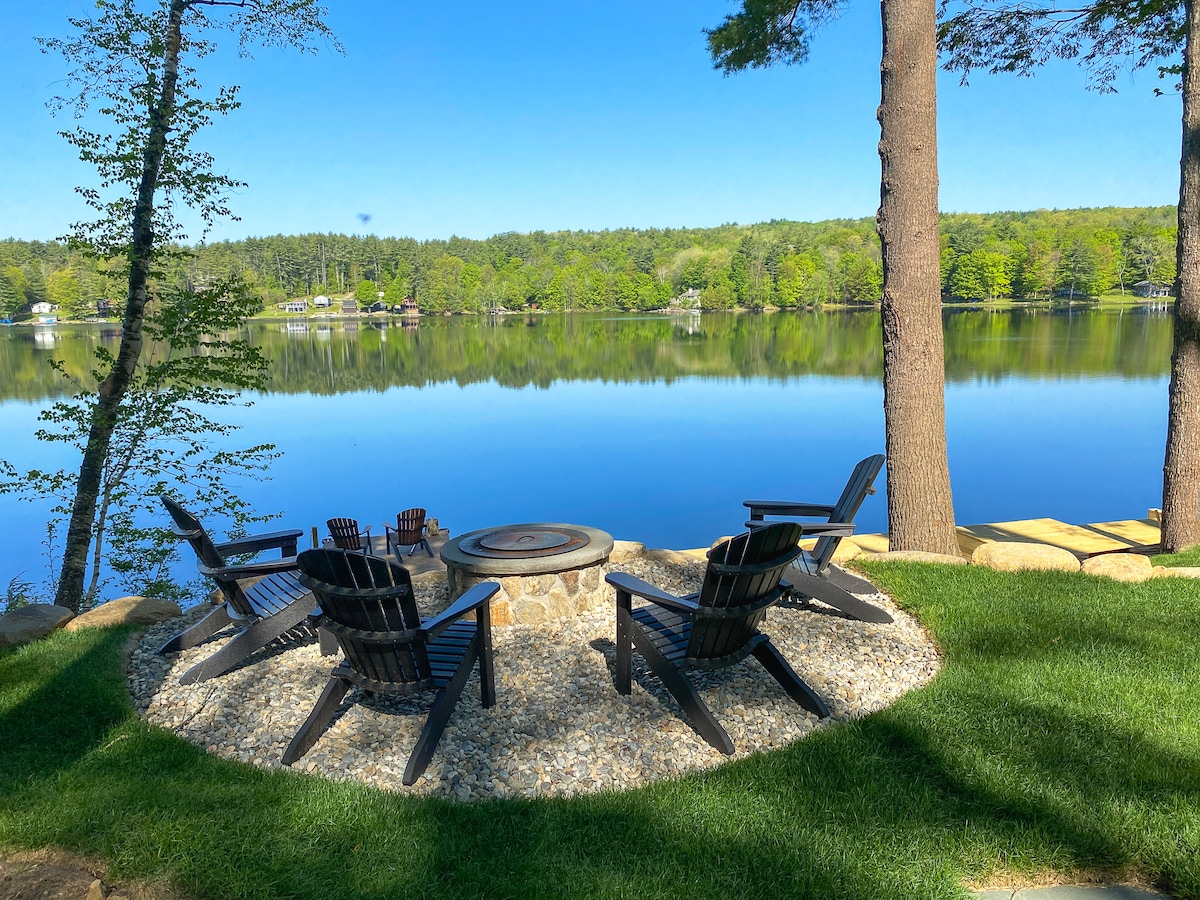 A serene outdoor seating area features four black Adirondack chairs arranged around a stone fire pit. The calm lake reflects the surrounding greenery, creating a peaceful atmosphere. Lush grass enhances the inviting space, ideal for relaxation by the water.
