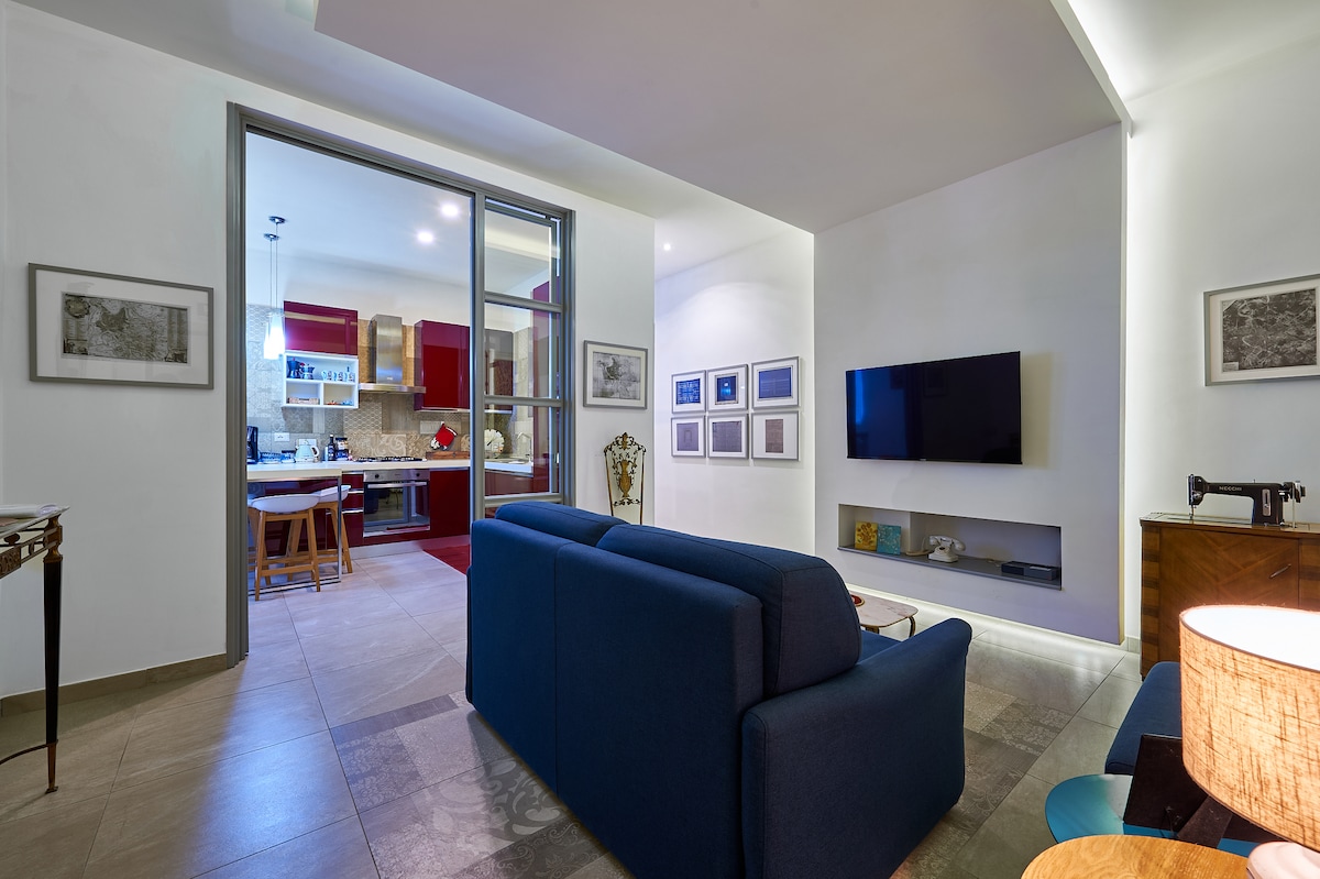 A modern living area features a dark blue sofa and a minimalist entertainment unit with a flat-screen TV. A dining area and kitchen are visible through large glass doors, showcasing red cabinetry. Framed artwork hangs on the walls, adding character to the space.