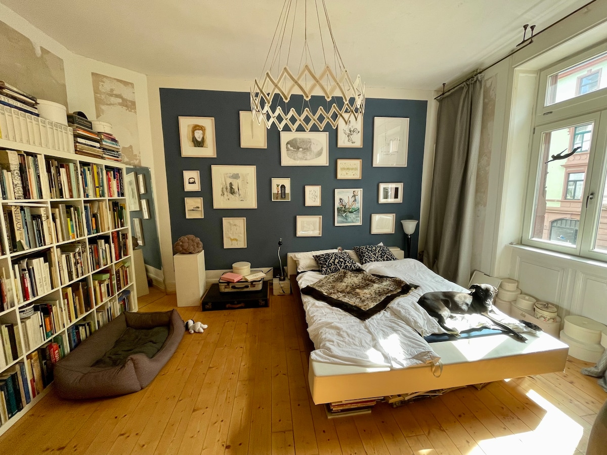A spacious bedroom features a large bed dressed in neutral linens with decorative pillows. An array of framed artwork lines the wall, creating an engaging focal point. A well-stocked bookshelf stands against one wall, while a cozy pet bed sits on the floor beside the bed.