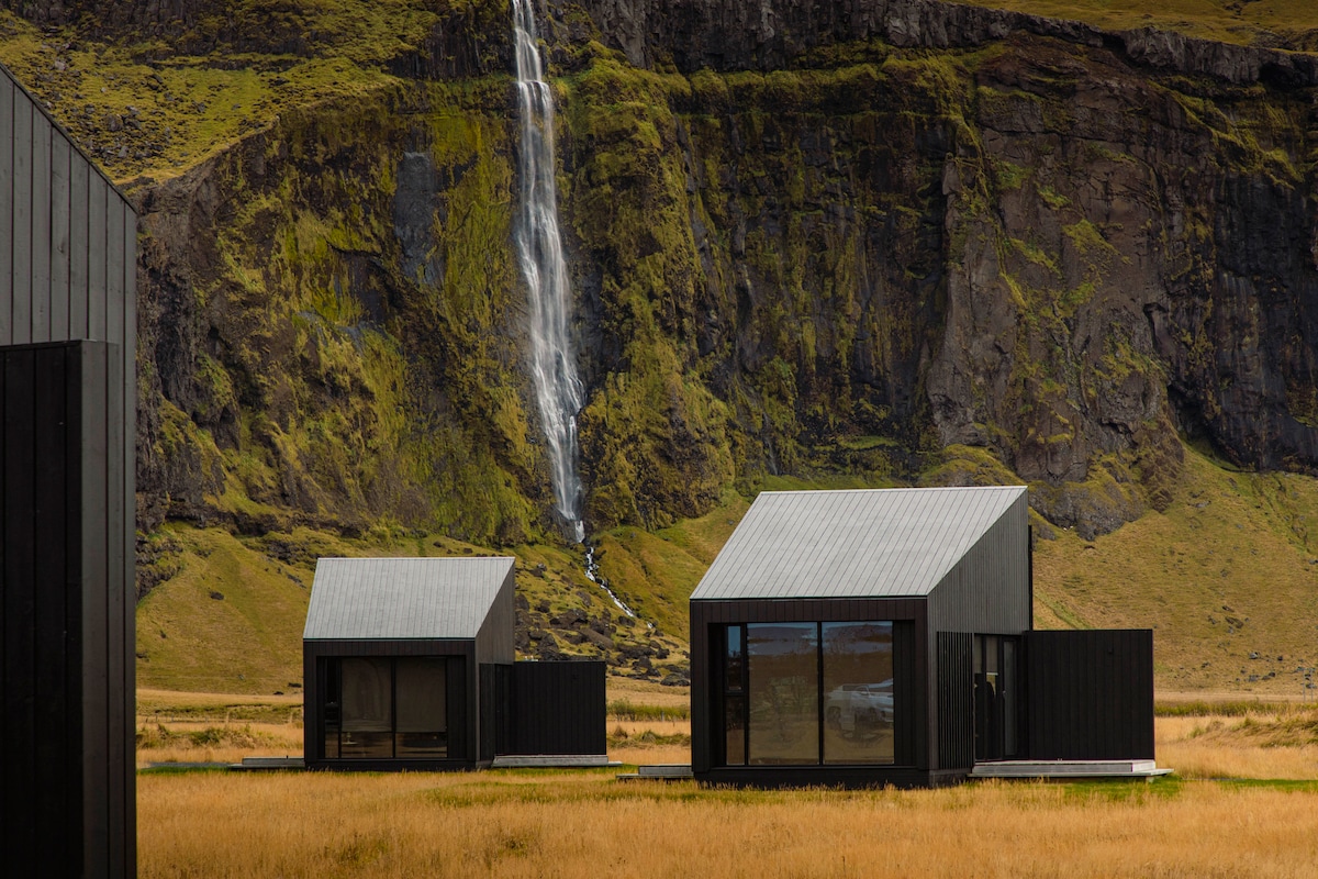 Two modern black cottages with angled roofs are set against a dramatic backdrop of a lush, green cliff and cascading waterfall. Vast grassy fields surround the cottages, enhancing the sense of tranquility in this natural setting.