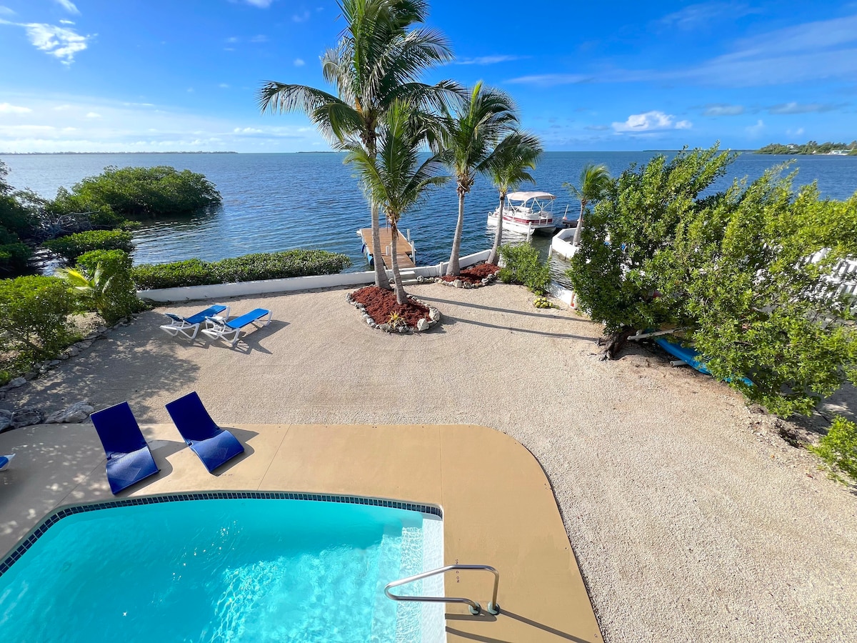 A private heated pool is showcased alongside a sandy area with lounge chairs, palm trees, and lush greenery. The waterway is visible in the background, with a small dock and boats moored nearby under a clear blue sky.