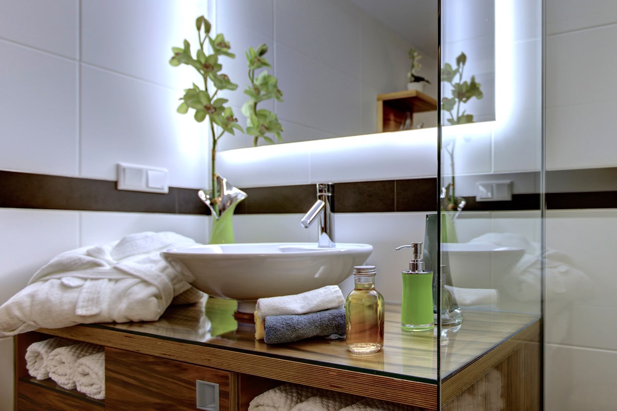 The modern bathroom features a stylish vessel sink on a wooden vanity. Soft towels are neatly arranged below, while green accents and an illuminated mirror add a fresh ambiance. Complimentary toiletries are displayed, highlighting a balanced combination of functionality and design.