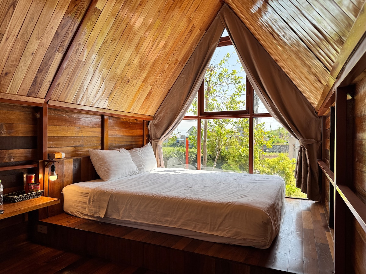 A cozy sleeping area is created with a spacious queen bed against a backdrop of wooden paneling. Large windows provide a view of the surrounding greenery, allowing natural light to fill the room, complemented by soft curtains.