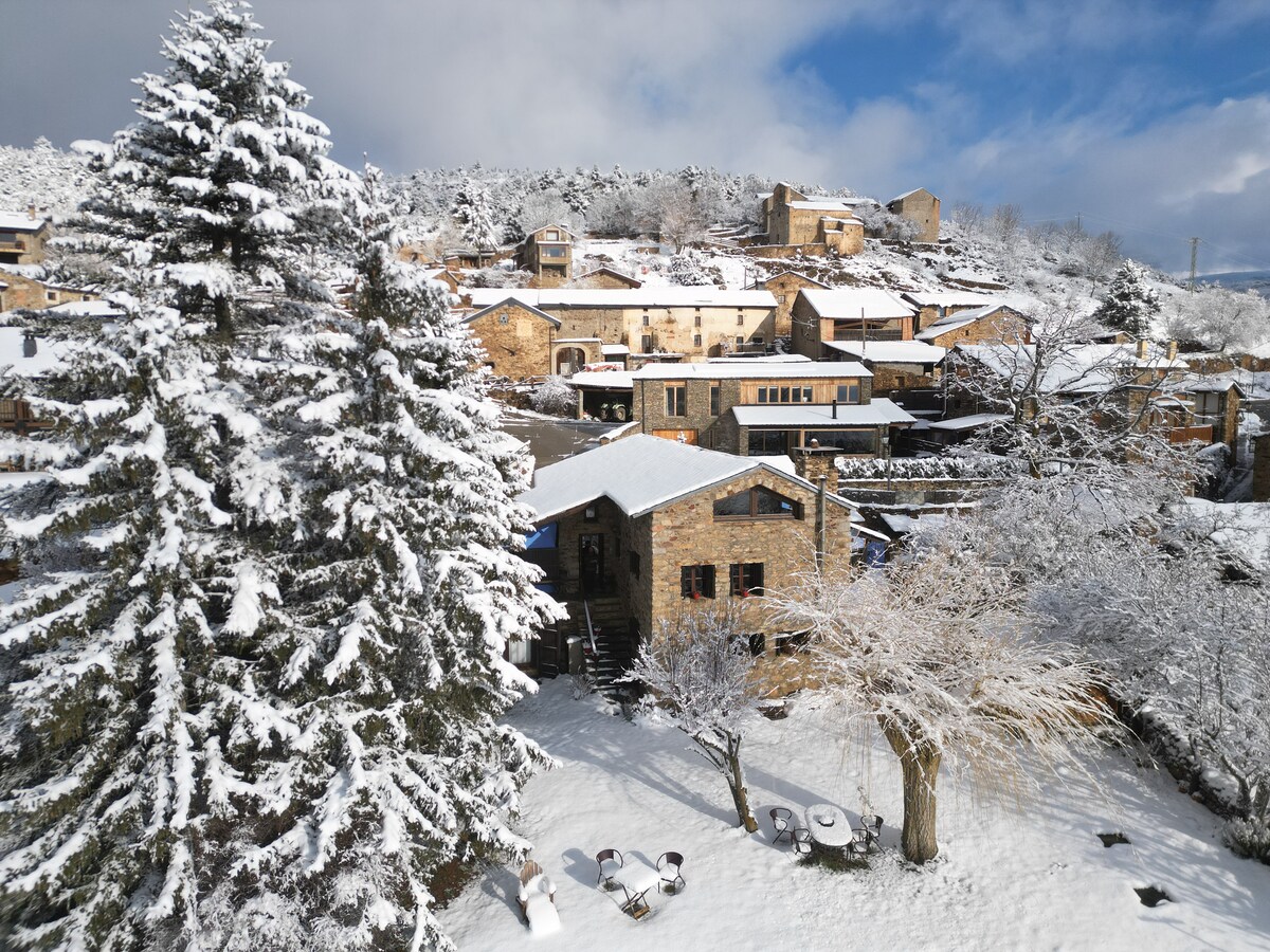 A traditional stone house is nestled among snow-covered trees and a serene landscape. The snow blankets the ground and rooftops, creating a picturesque winter setting. The image captures the essence of a tranquil getaway, framed by distant hills and clouds in a bright blue sky.