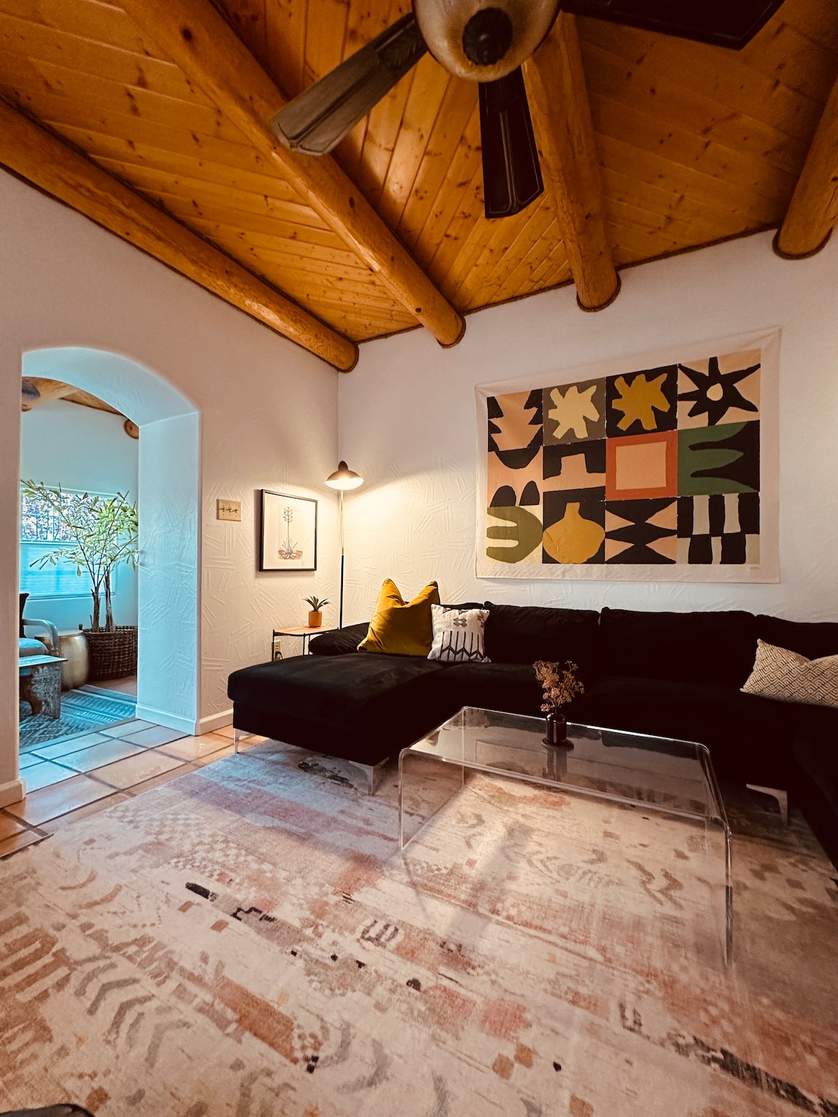 The living room features viga ceilings with a warm wooden finish. A black sectional sofa adorned with decorative pillows offers ample seating. A coffee table with a clear top sits atop a cozy area rug. Original artwork adds a touch of character to the space.