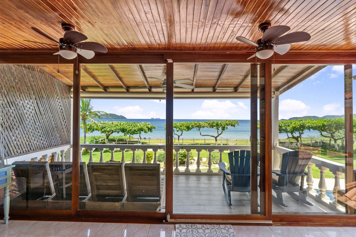 A covered balcony with a wooden ceiling features comfortable seating, including Adirondack chairs. Large sliding glass doors open to a stunning view of the beach and Pacific Ocean, with lush greenery lining the shore in the background.