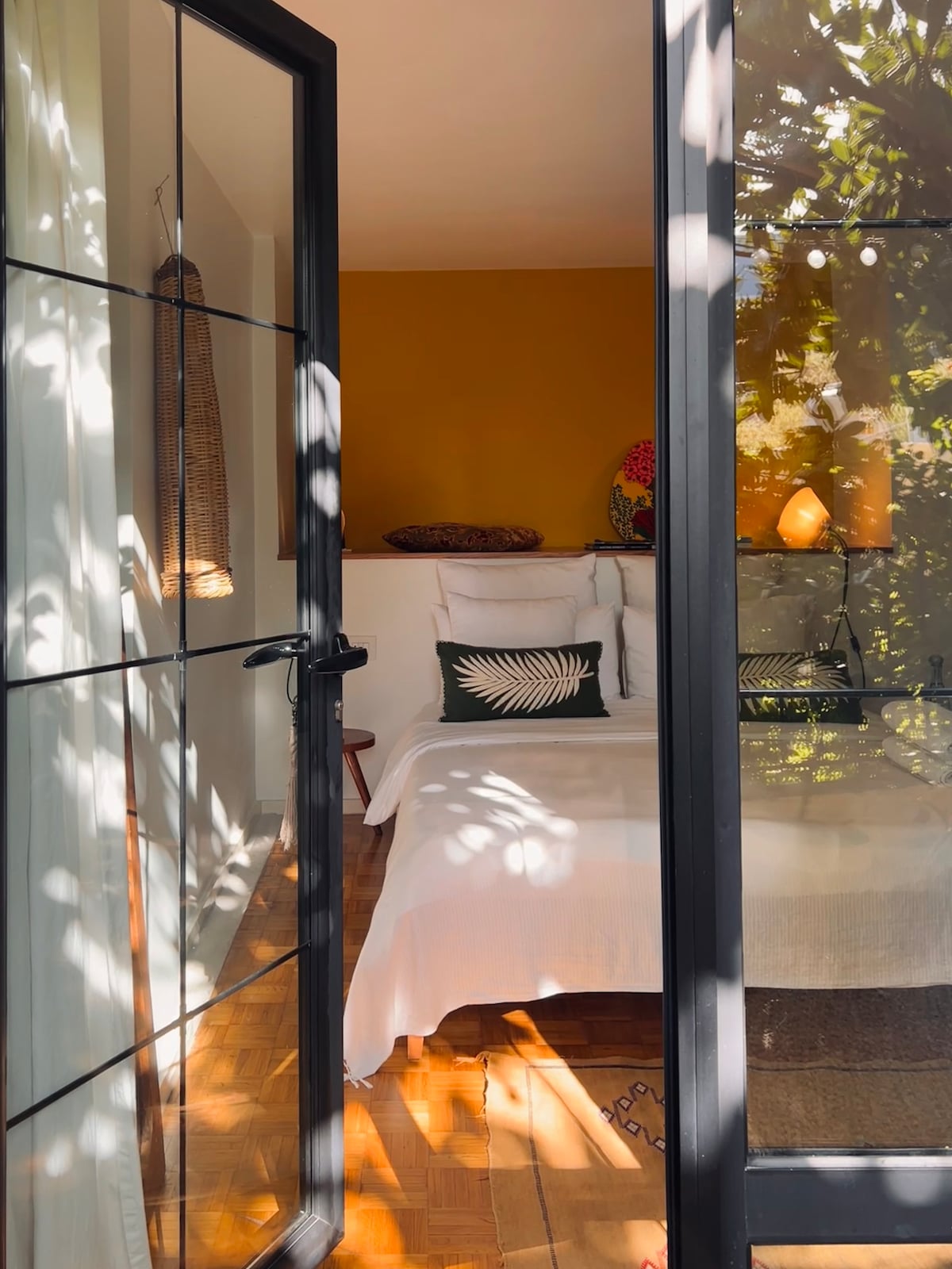 A guest suite is visible through glass doors, framed by shadows from nearby foliage. The space features a cozy bed adorned with decorative pillows and a warm accent wall. Natural light filters in, creating a serene and inviting atmosphere.