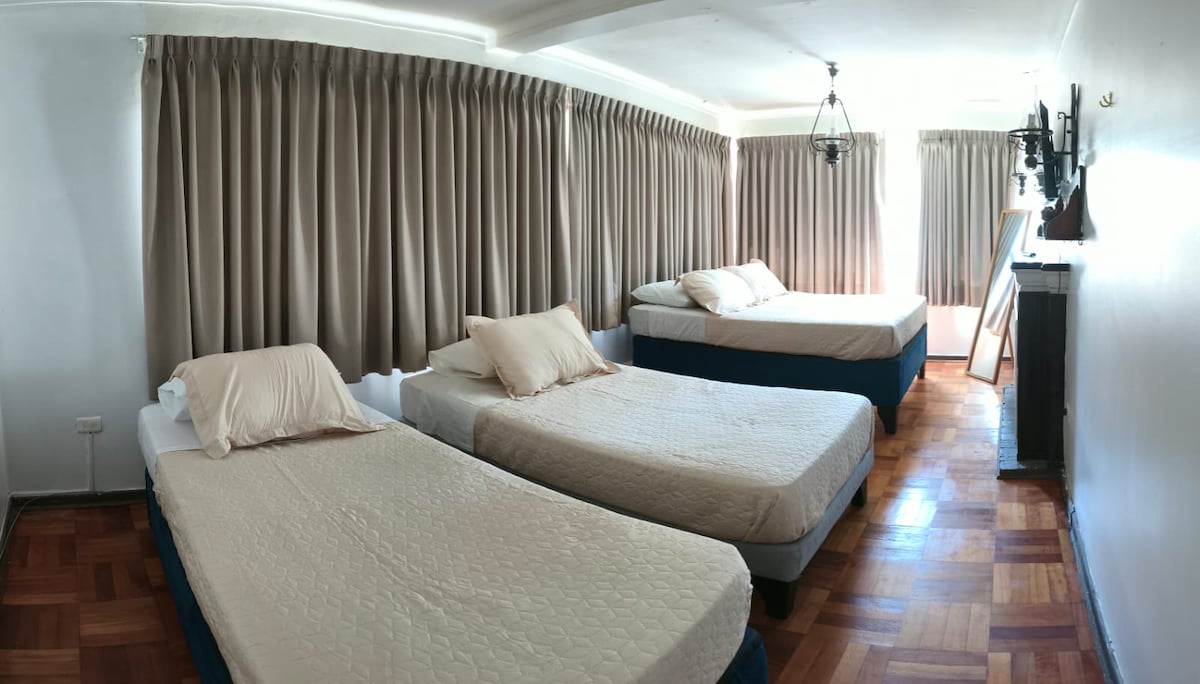 A spacious room features three individual beds with light-colored bedding. Soft curtains frame large windows, allowing natural light to fill the space. The polished wooden floor complements the warm tones, while a minimalist decor enhances the serene atmosphere.