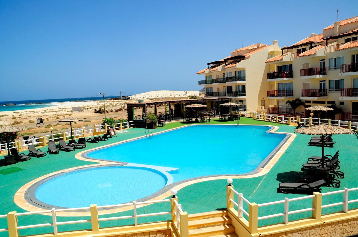 A spacious communal pool, surrounded by lounge chairs and umbrellas, invites relaxation. The pool area is bordered by a well-maintained green deck and offers a slight view of the nearby sea, enhancing the tranquil setting of the Vila Cabral 2 complex.