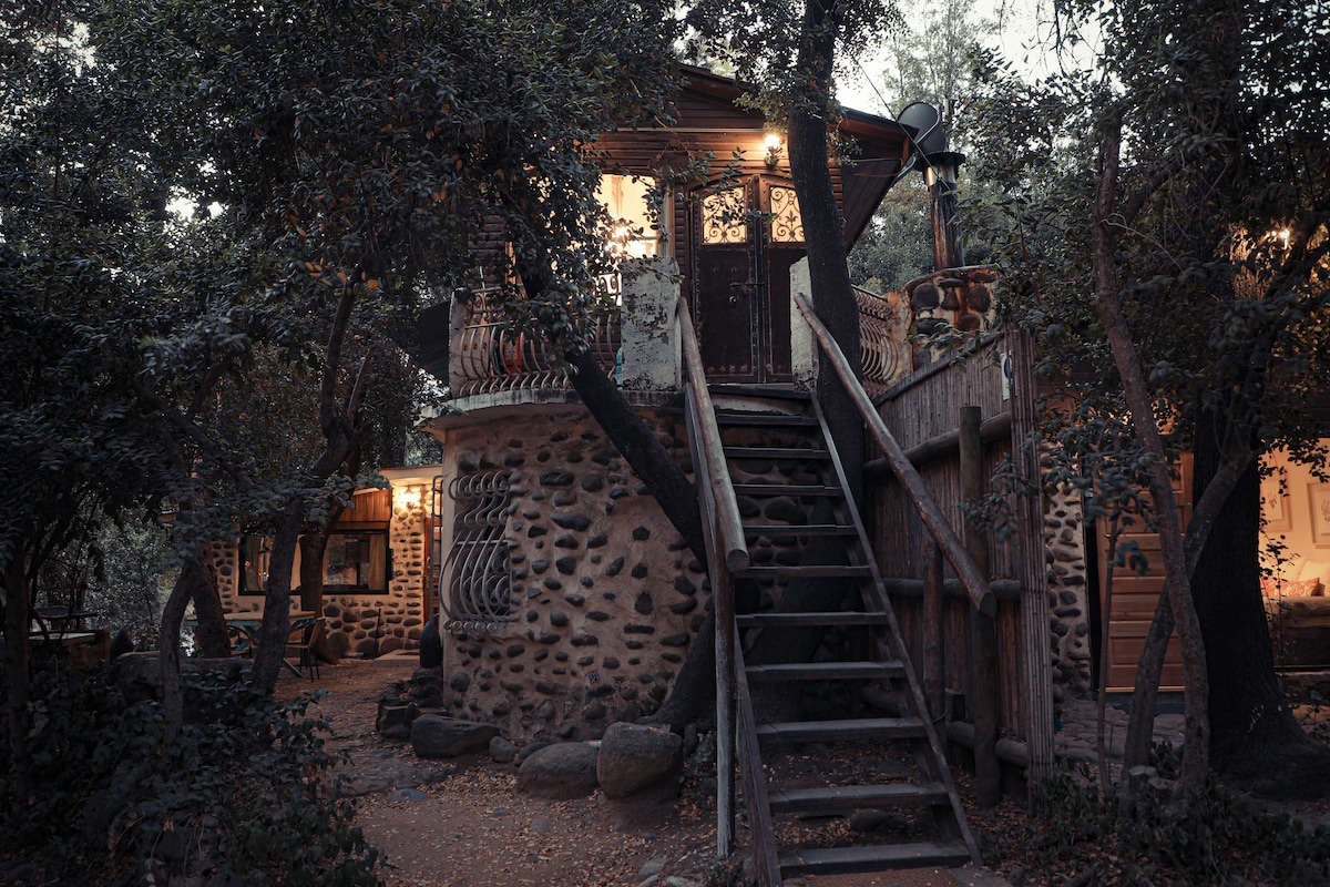 The entrance to the cabaña is framed by lush greenery, with a wooden staircase leading to an inviting door. The structure features natural stone accents and warm lighting, creating a cozy atmosphere amidst the surrounding trees.