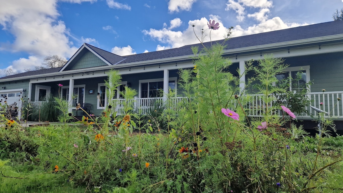 A charming home is framed by a variety of lively flowers and plants in the foreground. The porch features a white railing, while large windows allow natural light to brighten the interior. A clear blue sky with scattered clouds complements the inviting atmosphere.