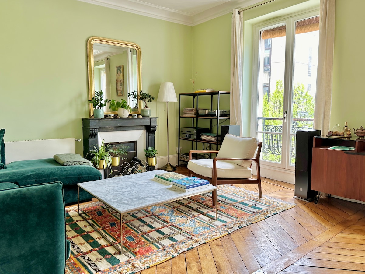 A light-filled living room features a green accent wall and hardwood flooring. A white marble coffee table sits on a colorful rug. A cozy armchair and a sectional sofa offer seating. A mirror reflects natural light, while a small balcony is visible through glass doors.