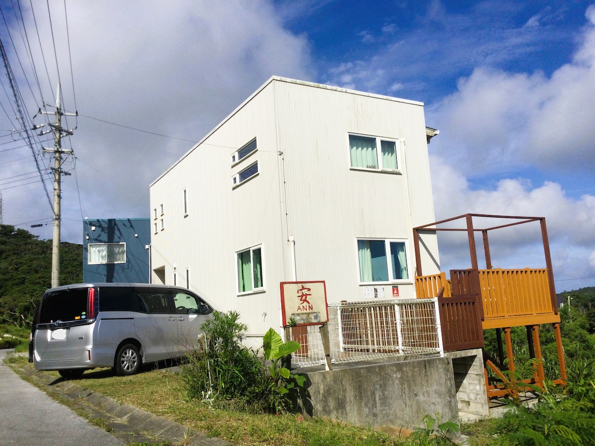 A two-story white condominium is situated on a hillside, displaying large windows and a modest balcony. A parked vehicle is positioned beside the building, while lush greenery surrounds the property. The cloudy sky adds a serene touch to the overall view.