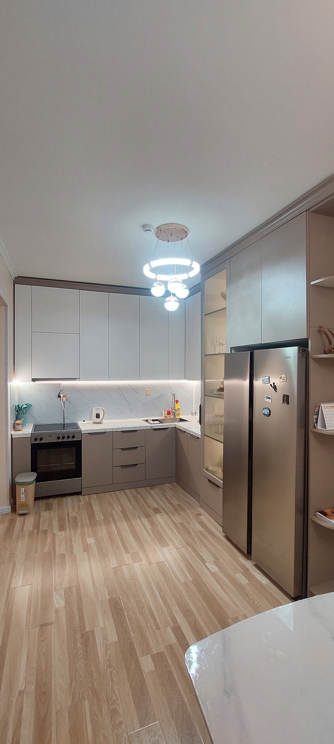 A modern kitchen space features light cabinetry with a contrasting darker accent. A sleek stainless steel refrigerator stands beside an oven and stovetop. Natural light illuminates the area, highlighting the clean lines of the cabinetry and the warm wooden floor, creating an inviting cooking environment.