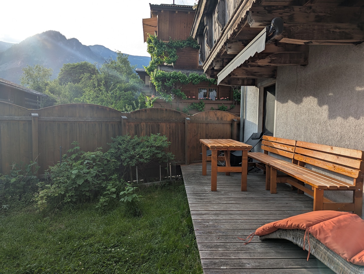 A private terrace is shown, featuring a wooden table and seating area made from warm-toned wood. Lush greenery surrounds the space, while mountains are visible in the background under a soothing light, enhancing the serene outdoor setting.