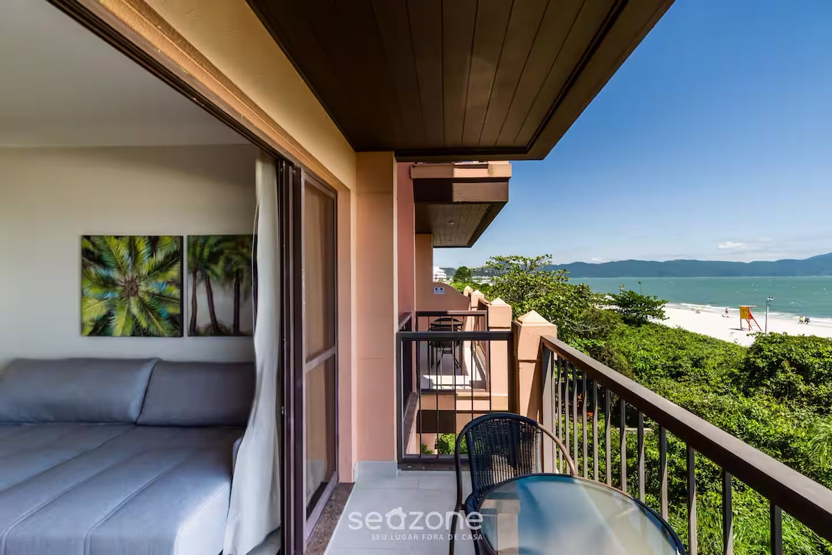 A private balcony features a table and chairs, offering a panoramic view of the beach and surrounding greenery. A sliding door leads to the interior space, which includes a comfortable sofa, creating an inviting area for relaxation.