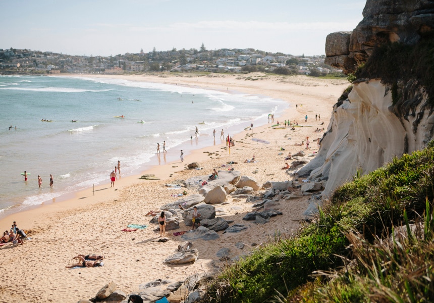 A sandy beach stretches into the distance, bordered by gentle waves lapping at the shore. Visitors engage in various activities, while others relax on towels spread across the sand. Coastal cliffs provide a natural backdrop, with patches of greenery visible along the edge.