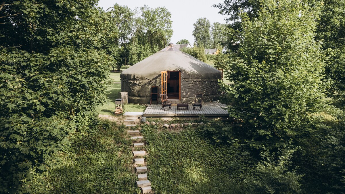 A yurt, constructed from natural materials, is nestled among lush greenery. Wooden steps lead up to a spacious terrace with seating, framed by trees. Large, open doors reveal the inviting atmosphere inside the yurt.