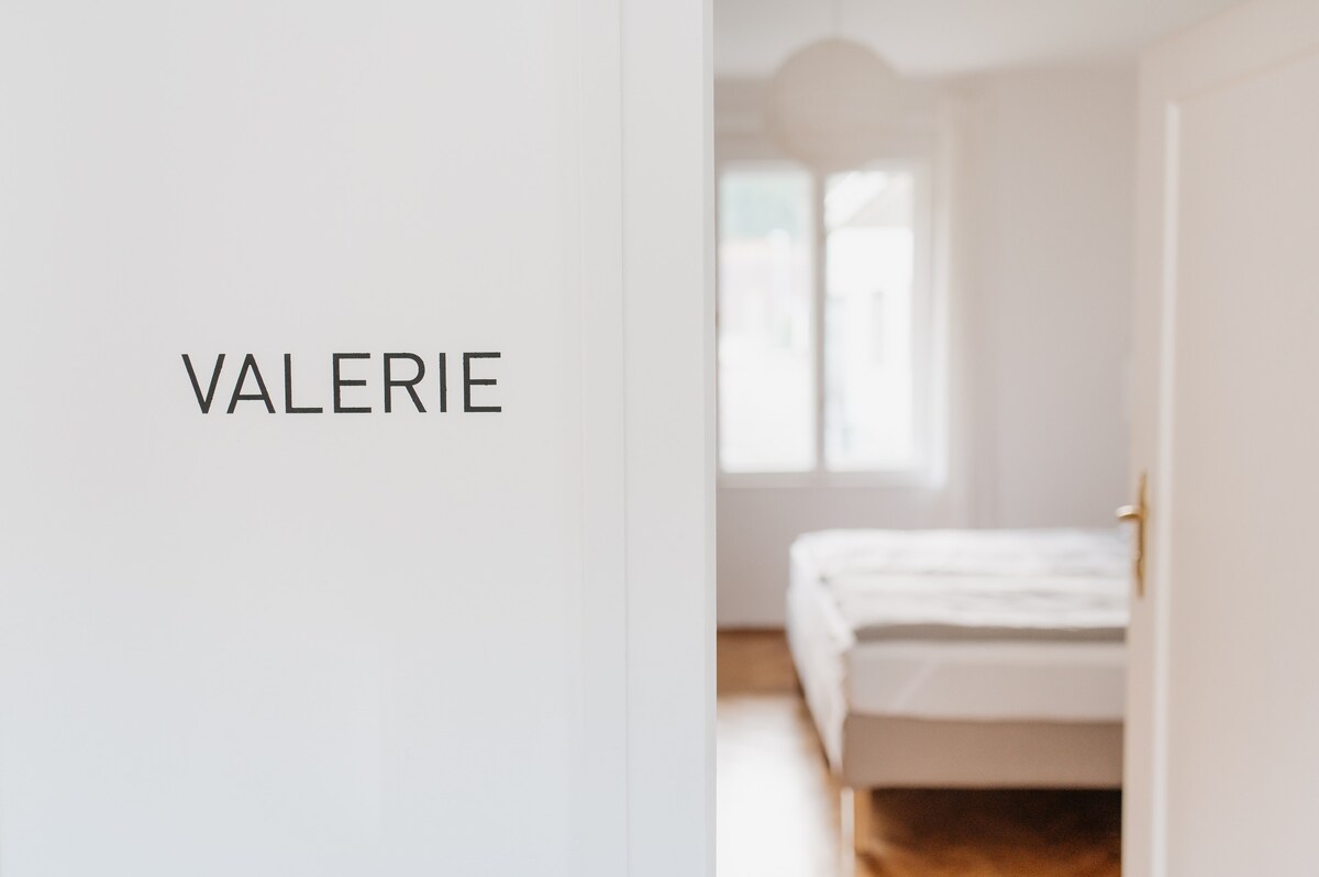 A door labeled 'VALERIE' opens into a softly lit room featuring a spacious bed with an unmade mattress. Natural light fills the space through a nearby window, enhancing the subtle color palette of the room, and wooden flooring complements the overall design.