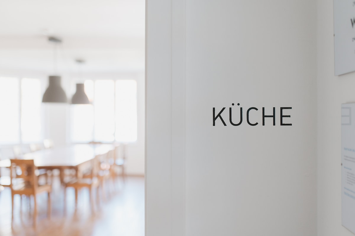 The image shows a spacious kitchen area visible through an open doorway. A large dining table surrounded by wooden chairs occupies the sunlit room, with multiple windows allowing natural light to fill the space. Pendant lights hang above the table, enhancing the inviting environment.