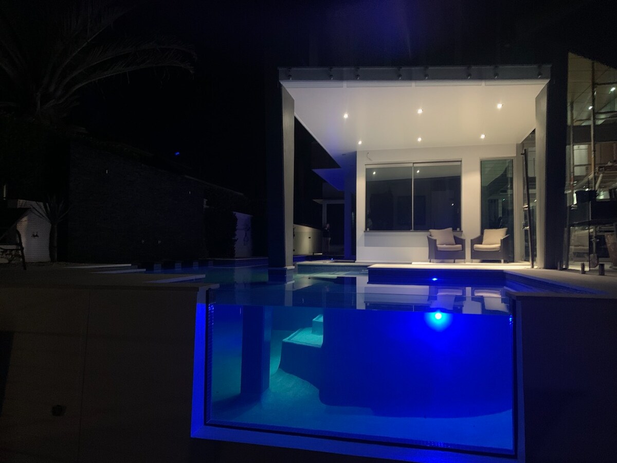 The image showcases a modern guest suite illuminated at night, with large windows revealing a lounge area and two chairs. A tranquil pool reflects a soft blue light, enhancing the serene atmosphere. Palm trees are visible, adding to the outdoor setting.