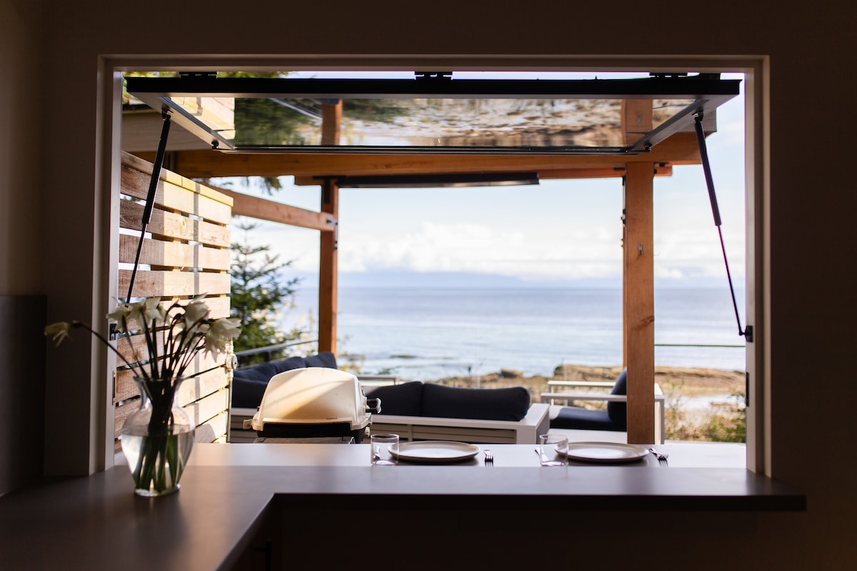 An open window reveals a panoramic view of the ocean, with outdoor seating visible on the deck. A clean countertop holds two place settings of plates and utensils. The scene conveys a connection between indoor and outdoor spaces, framed by natural wood beams.