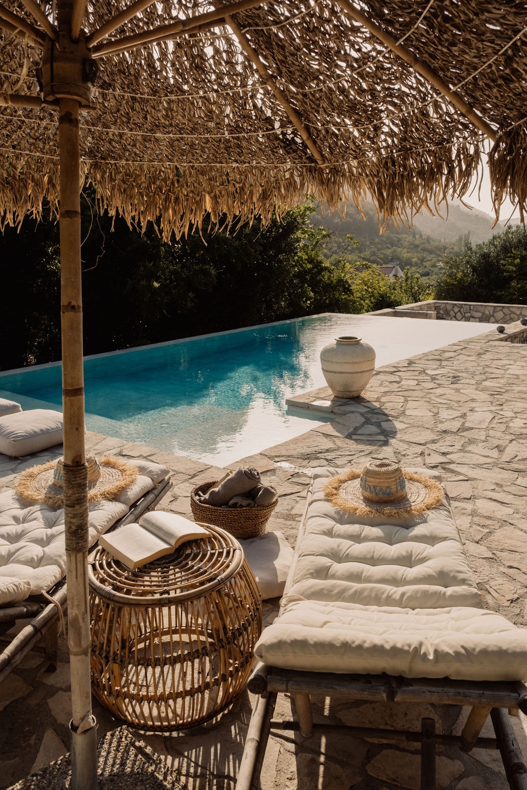 A sunlit terrace features comfortable lounge chairs positioned near a heated infinity saltwater pool. A thatched umbrella provides shade, while a woven basket and a decorative pot add to the natural ambiance. Lush greenery surrounds the area, creating a serene outdoor space.
